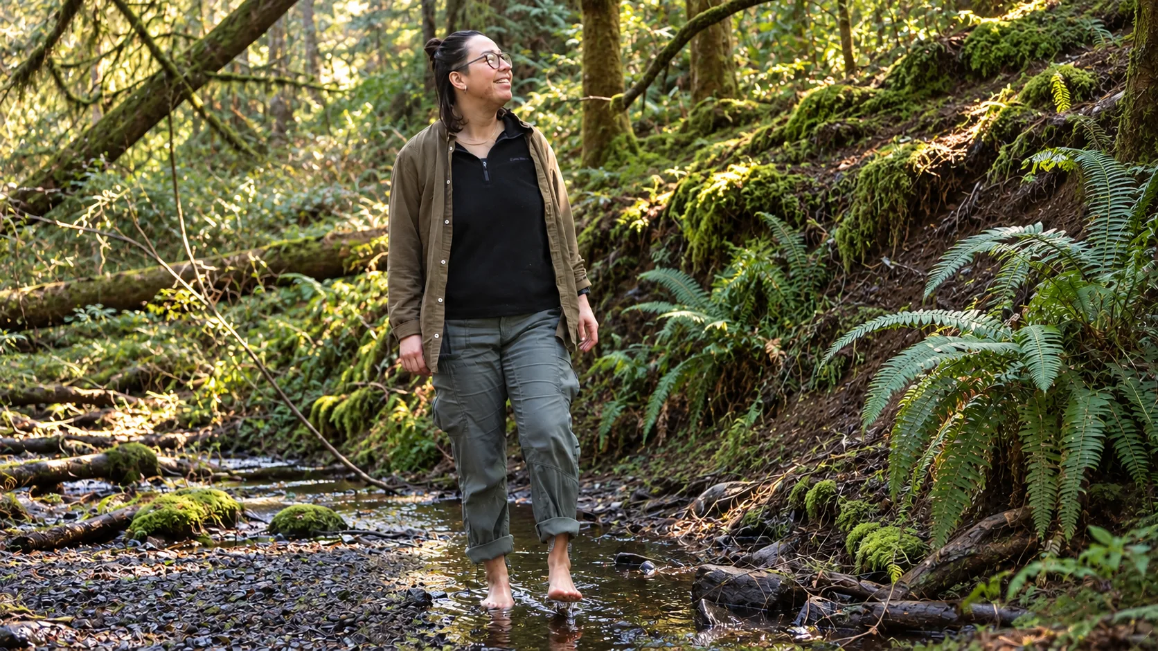 Woman walking barefoot through a shallow forest creek surrounded by moss-covered logs and sword ferns