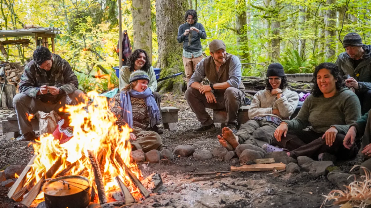 Wild Path students and mentors laughing together around a campfire in a mossy forest camp
